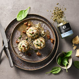 Drei Knödel mit Kräutern und Käse auf Teller, garniert mit Nüssen, daneben Gewürzglas und frische Zutaten.